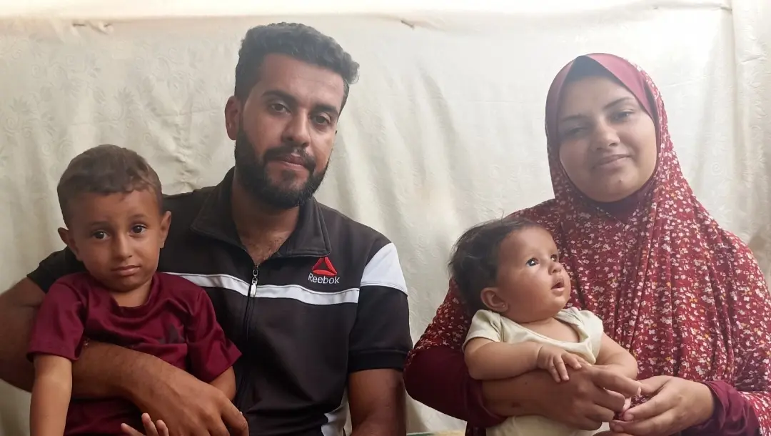 Mohammed holding Ibrahim at Welcome to Gaza sign with destroyed buildings in background